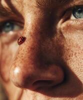 A highly detailed macro portrait of a persons face with light freckles, focusing on the nose where a red ladybug with black spots is gently crawling. photo