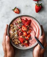 A delicious thick strawberry smoothie bowl topped with fresh sliced strawberries, crunchy granola, and black chia seeds. The bowl is held in two hands photo