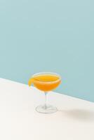 A minimalist orange cocktail served in a classic coupe glass with a sugar rim and a twist of orange peel as garnish, placed on a white surface against a pastel blue background. photo