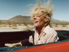 A joyful young blonde woman with wind-blown hair, laughing while riding in a red convertible through a desert landscape. She wears a striped button-up shirt, and her hair is messy from the speed. photo