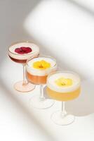 Three cocktails with foam, each topped with an edible flower, served in elegant pastel glasses with varied shapes one with a gold rim, one with scalloped edges, and one ribbed. photo