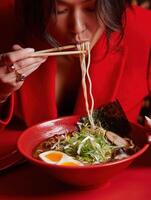 A woman in a bright red blazer is eating ramen, lifting long noodles with sprouts using chopsticks photo