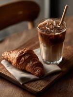 A crispy croissant and a glass of iced latte with a straw served on a wooden board with a linen napkin. photo