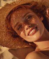 A red-haired woman lies smiling on the sand, holding a straw hat that casts a patterned shadow on her face. photo