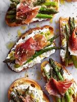 Realistic food photography of open-faced sandwiches made with rustic toasted bread, creamy ricotta spread, roasted asparagus spears, and thin slices of crispy prosciutto on top. photo