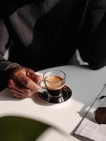 A mans hand holds a glass cup of espresso on a minimalist white table. photo
