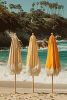 Three closed vintage-style beach umbrellas with fringe details, standing on a sunny tropical beach with turquoise waves in the background and palm trees in the distance. photo