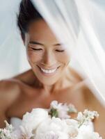 Close-up portrait of a smiling bride in a strapless white dress, holding a soft pastel bouquet in front of her and gently lowering her veil over her face. photo