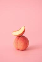 Minimalist food composition showing a whole ripe peach with a fresh slice balanced on top, placed against a soft matte pink background photo