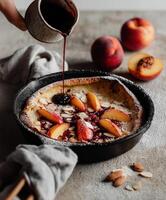 A rustic cast iron skillet with a freshly baked Dutch baby pancake, topped with sliced ripe peaches, almond flakes, and a dusting of powdered sugar, while a hand pours dark berry syrup over the top. photo