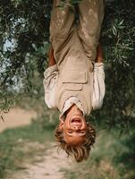 A joyful young boy with tousled blond hair hanging upside down from a tree branch, laughing with wide open mouth, wearing vintage beige corduroy overalls and a white shirt. photo