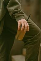 A close-up of a person in an olive green outfit, holding a plastic cup of iced tea with a black straw. photo