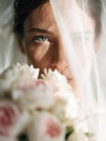 A bride in a white dress holding a delicate pastel bouquet of roses, partially hidden behind a transparent veil. Close-up shot, focus on her eye and face through the sheer fabric. photo