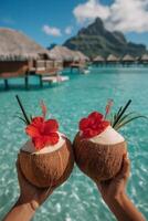 A pair of hands holding two fresh coconuts, each topped with a vivid red hibiscus flower, black straw, and a tropical plant leaf garnish photo