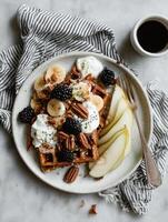 A round white ceramic plate with crisp golden waffles topped with banana slices, blackberries, pecans, chia seeds, and dollops of yogurt. photo
