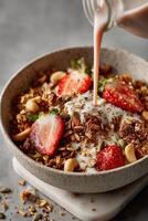 A white ceramic bowl filled with granola, fresh strawberry slices, cashews, and sunflower seeds. A stream of creamy pink nut milk is being poured from a glass bottle into the bowl. photo