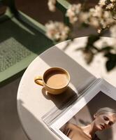 Minimalist flat-lay of a yellow coffee cup and an open fashion magazine on a round beige table, with soft sunlight and flower shadows creating a peaceful mood. photo