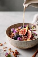 A speckled ceramic bowl filled with granola, halved fresh figs, blackberries, and edible purple flowers. A stream of creamy nut milk is being poured into the bowl from above, creating a dynamic flow. photo