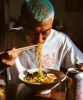 A man with turquoise hair and a white T-shirt featuring a Japanese print is leaning over a bowl, slurping ramen noodles intensely. photo