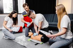 Young man instructor helping to make first aid heart compressions with dummy during the group training in room photo