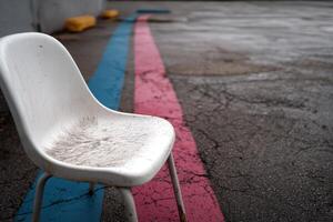 White chair placed on cracked pavement beside blue and pink lines near an empty parking area photo