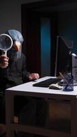 vertical mascarado hacker usando ampliação vidro enquanto trabalhando em computador. espião golubiro video