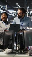 Vertical Smiling engineering team in server room reviewing infrastructure diagnostics on devices. IT professionals in data center analyzing performance data to maintain system integrity and uptime, camera A video
