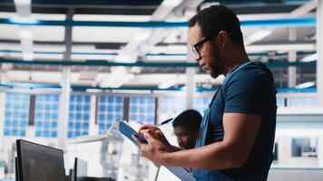 Black engineer in photovoltaics factory reviews performance of solar panels assembly lines, checking paperwork for finetuning. Supervisor inspecting solar cell machinery. Camera A. video