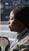 Vertical Data center system administrator using PC to monitor neural network AI visualization. African american woman in server room using computer to oversee rigs powering machine learning processes, camera A video