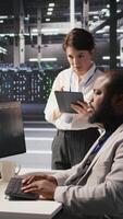 Vertical Server room manager supervise technician examining deep learning algorithms. Data center team leader and African american employee review performance dashboards of AI equipment rigs, camera A video