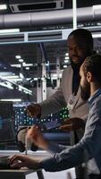 Vertical Data center engineers use laptop, collaborating on doing AI software updates. Server room teamworking colleagues doing brainstorming, optimizing infrastructure using artificial intelligence, camera A video