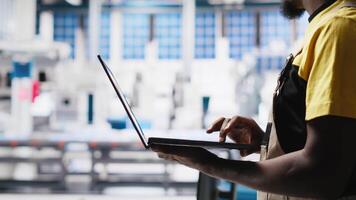 Black technician in solar panel facility reviews photovoltaic system performance on laptop. Specialist analyzing production line metrics to optimize efficiency in industrial plant. Camera A. video