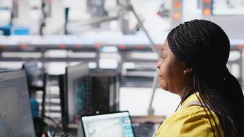 Black factory employee oversees solar system performance at desk seated beside production line machines. Female technician analyzes solar energy flow and updates system software. Camera A. video