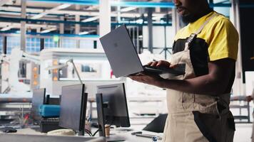 Black engineer in photovoltaics factory using laptop to diagnose malfunctioning systems, debugging. Solar panels manufacturing plant worker reduces downtime, troubleshooting. Camera B. video