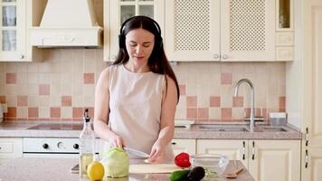 a Woman in kitchen wearing headphones, slicing cucumber, snacking and enjoying music while making salad video