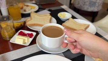 good coffee in a white cup for breakfast, a woman's hand holds it, side view, in the background lie toasts with butter, jam and other table setting, buffet hotel all inclusive, caffeine enjoyment video