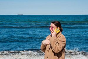 Young caucasian female at the beach wearing a brown coat, talking on a yellow phone with purple sunglasses, stands by the ocean on a clear day. Digital nomad lifestyle. Remote work photo