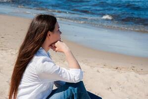 Young caucasian female in white shirt and jeans sits thoughtfully on a sandy beach looking across waves under a clear blue sky embracing tranquility. Digital nomad lifestyle. Remote work photo