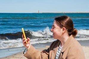 Young caucasian female enjoying a relaxing beach day, smiling as she takes a selfie with her phone against a backdrop of waves and a distant lighthouse. Digital nomad lifestyle. Remote work photo