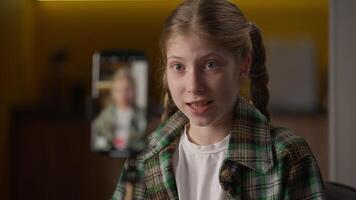 Cute cheerful Caucasian schoolgirl little girl teenager kid child influencer blogger talking online streaming blogging speak emotion record mobile phone camera on tripod smartphone blog at home video