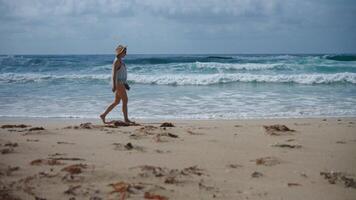 A woman strolls along a sandy beach, her bare feet sinking into the soft shore as waves crash gently in the background. Dressed in a fringed beach cover-up and a straw hat video