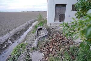 Old wooden boat abandoned on dry canal bank next to white building with rusty door and green vegetation photo