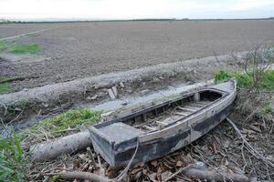 Abandoned old wooden boat lying in dry canal surrounded by grass mud and farmland under cloudy sky photo