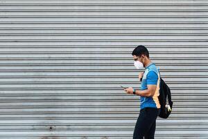 Hispanic male with a protective face mask and a backpack looking at his phone in front of a garage. photo