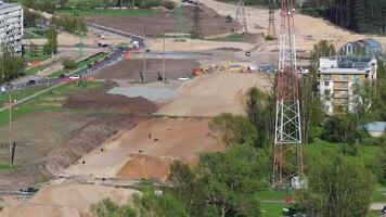 Active construction site with trucks, excavators, and equipment in motion. Features a red and white tower, power lines, forest, and urban buildings. video