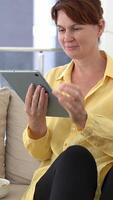Relaxed atmosphere envelops middle-aged woman on couch, casually enjoying junk food while immersed in digital content on tablet. Comfortable outfit, leisure vibe, blending technology with comfort video