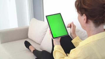 Woman with Red Hair in Yellow Shirt Relaxing on Sofa, Holding Tablet with Green Chromakey Screen, Touching Display, Browsing Online, Digital Connection, Smart Technology in Bright Home Setting video
