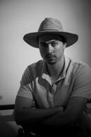 Man in a straw hat radiates calm confidence in monochrome setting while sitting in a casual pose photo