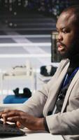 Vertical Stressed technician in data center managing AI server infrastructure, drinking coffee. African american worker using PC to run artificial intelligence, panicked by deadline, camera A video