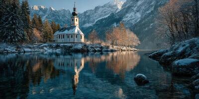 Snow-covered church reflected in calm lake with mountains in background during winter morning photo